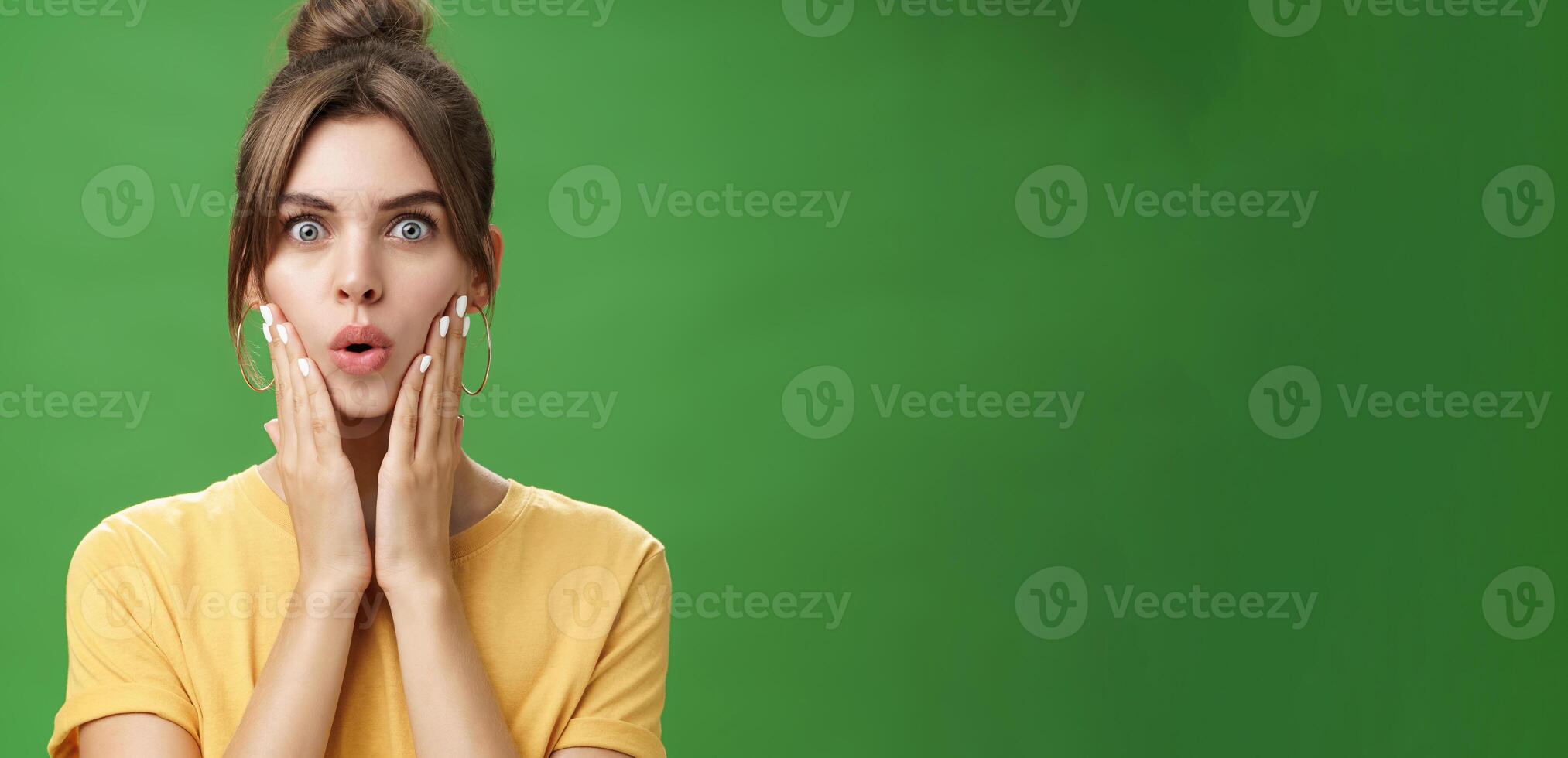 Portrait of impressed and shocked cute feminine woman in yellow t-shirt folding lips from excitement and interest touching cheeks surprised reacting to amazing rumor posing over green background photo
