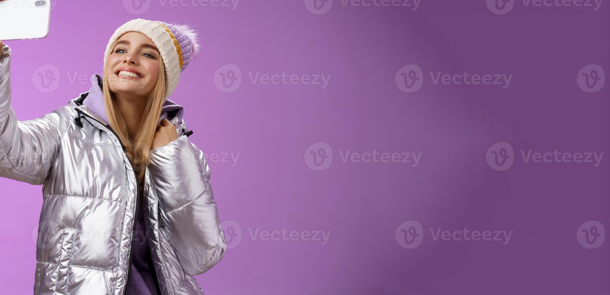 Happy delighted carefree fair-haired charming european woman in silver winter jacket hat raising smartphone horizontally taking selfie smiling mobile phone display, purple background photo