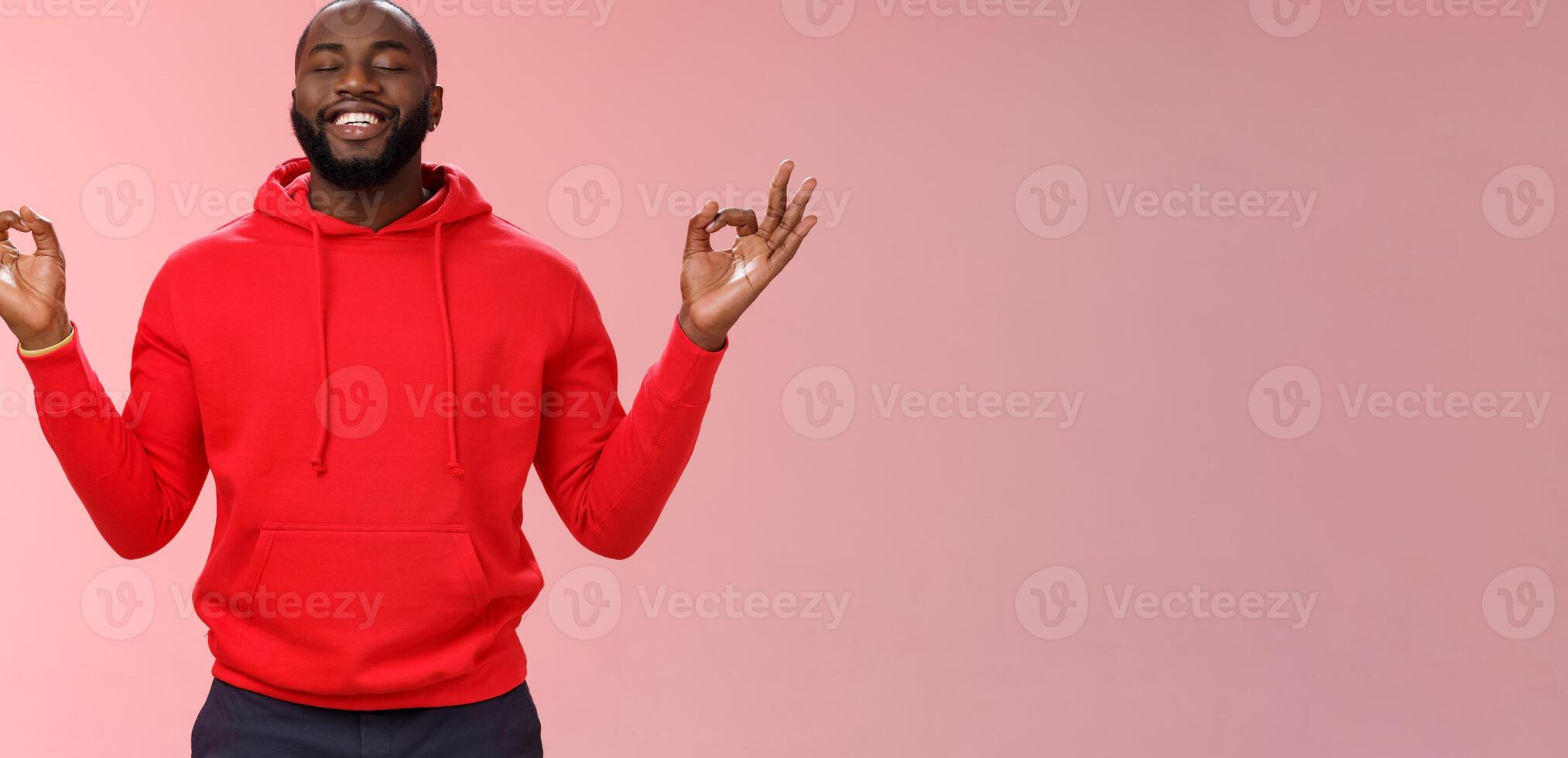 Happy african american bearded man in red hoodie meditating found peace nirvana smiling delighted close eyes relaxed relieved standing lotus mudra pose search zen, practice yoga, pink background photo