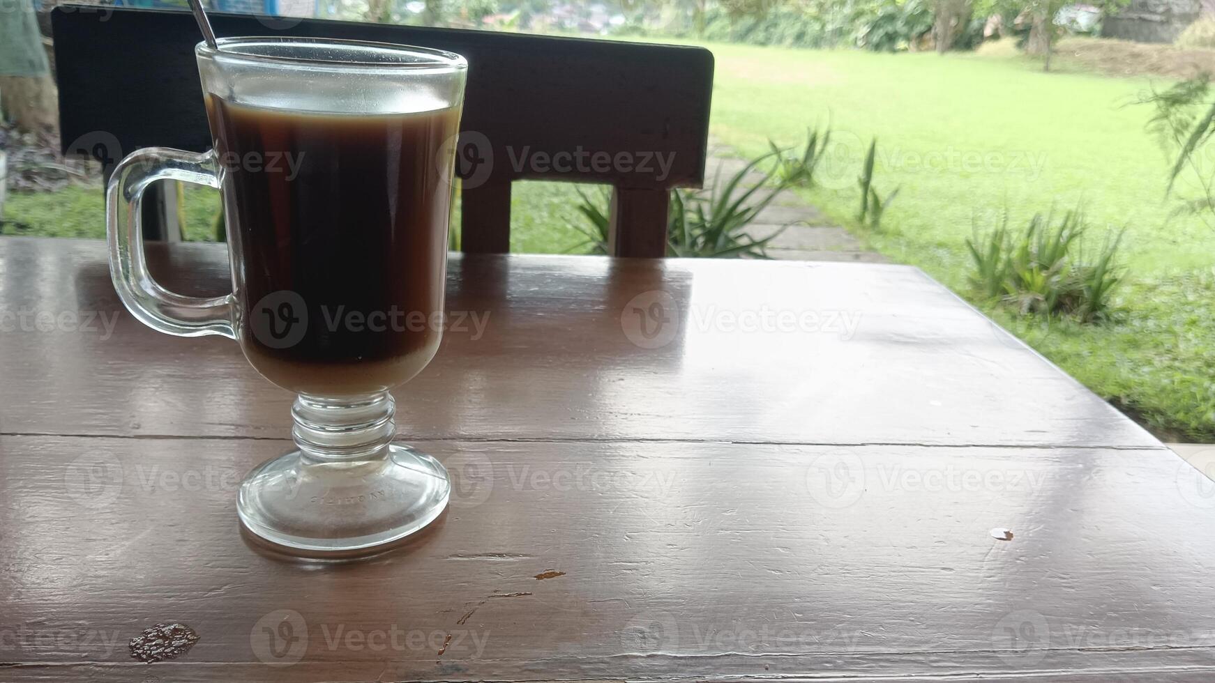 A glass of coffee with creamer on a wooden table outdoors with a green lawn in the background. photo