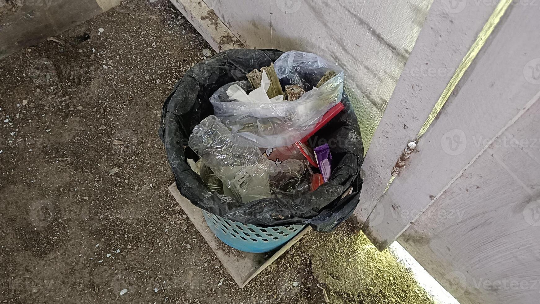 A black trash bag overflowing with garbage, including plastic bottles, plastic bags, and other assorted trash, sits in a blue trash can against a white wooden fence. photo