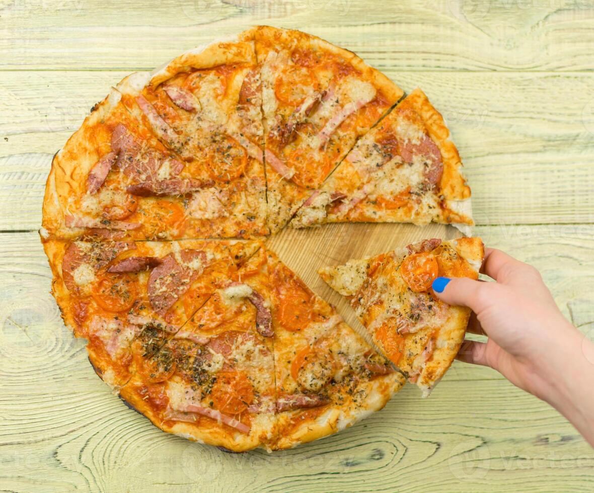 Freshly prepared homemade pizza on a wooden background. A woman's hand takes a piece of pizza. photo