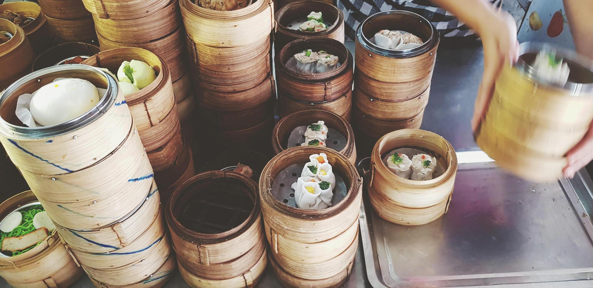 Many kind of hot Dim Sum in weave bamboo basket on stainless steel table for sale and movement of hand taking food for serving customer in Chinese restaurant. Focus on Food and Service concept. photo
