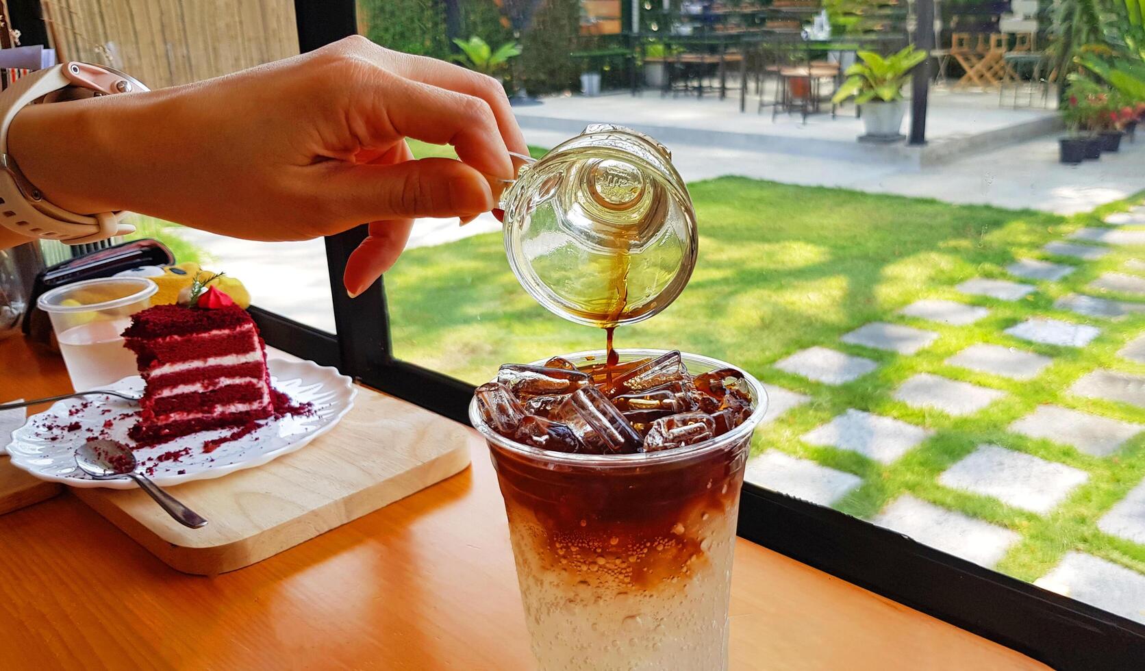 Hand of people pouring short of dark espresso coffee or tea into glass of ice soda with sweet red cake and green grass background. Refreshment drinking and Mixing food concept. photo