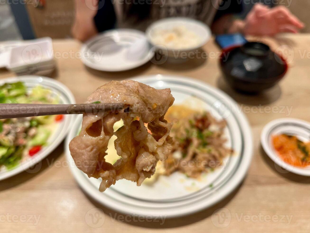 chopsticks holding a slice of tender, cooked meat with a blurred background of a Japanese meal. The focus is on the texture and juiciness of the meat, showcasing the delicious photo