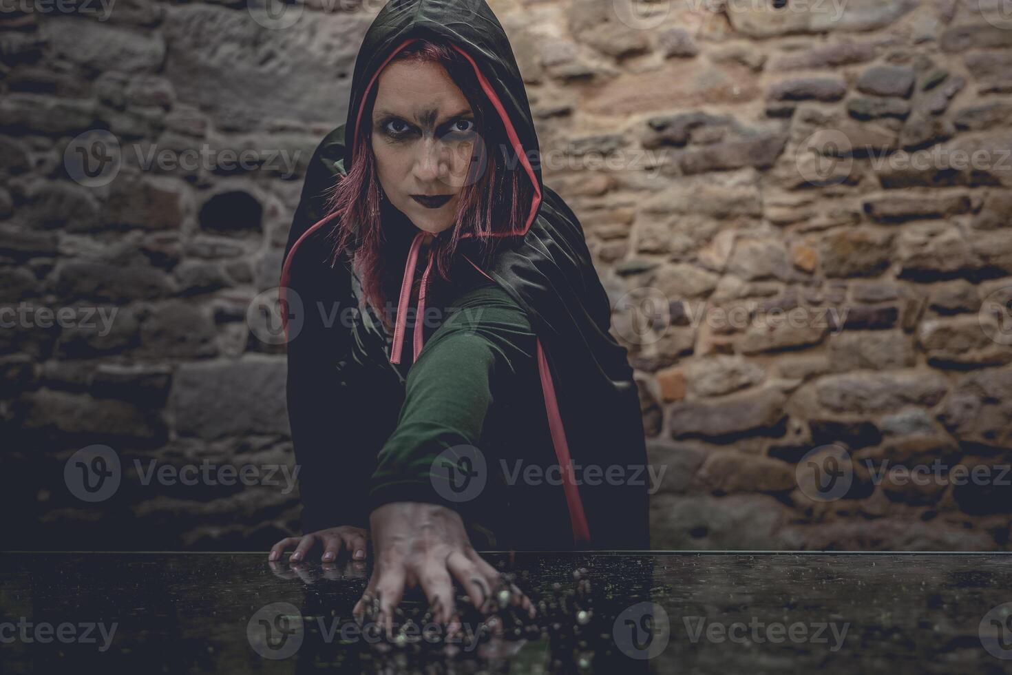 woman in a black cloak with red hair and black makeup is reaching for something on a table photo