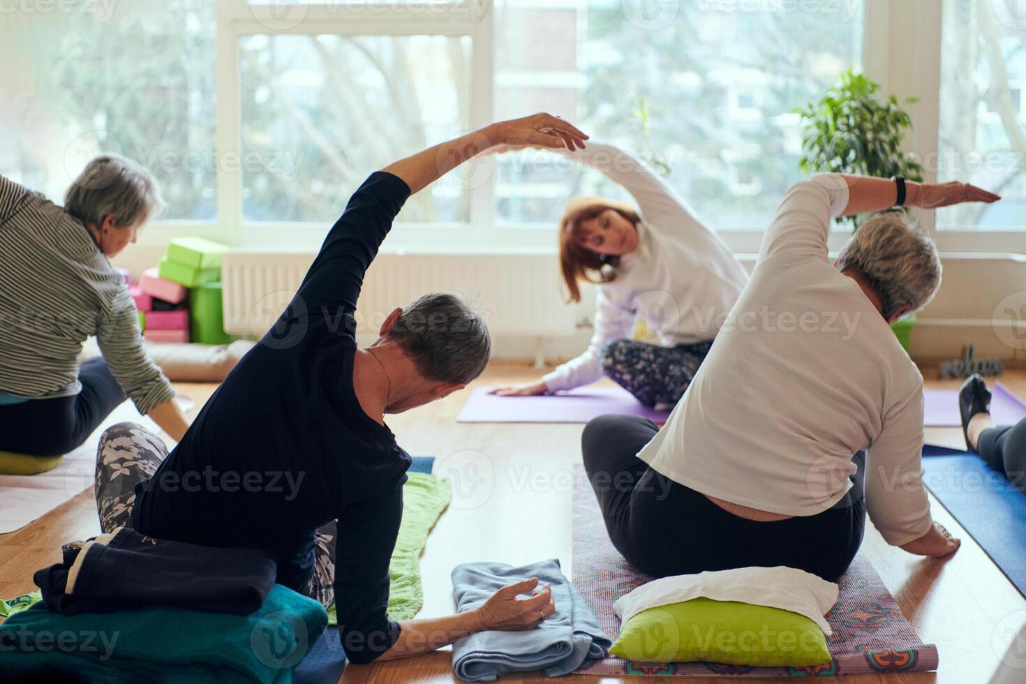 A group of senior women engage in various yoga exercises, including neck, back, and leg stretches, under the guidance of a trainer in a sunlit space, promoting well-being and harmony photo