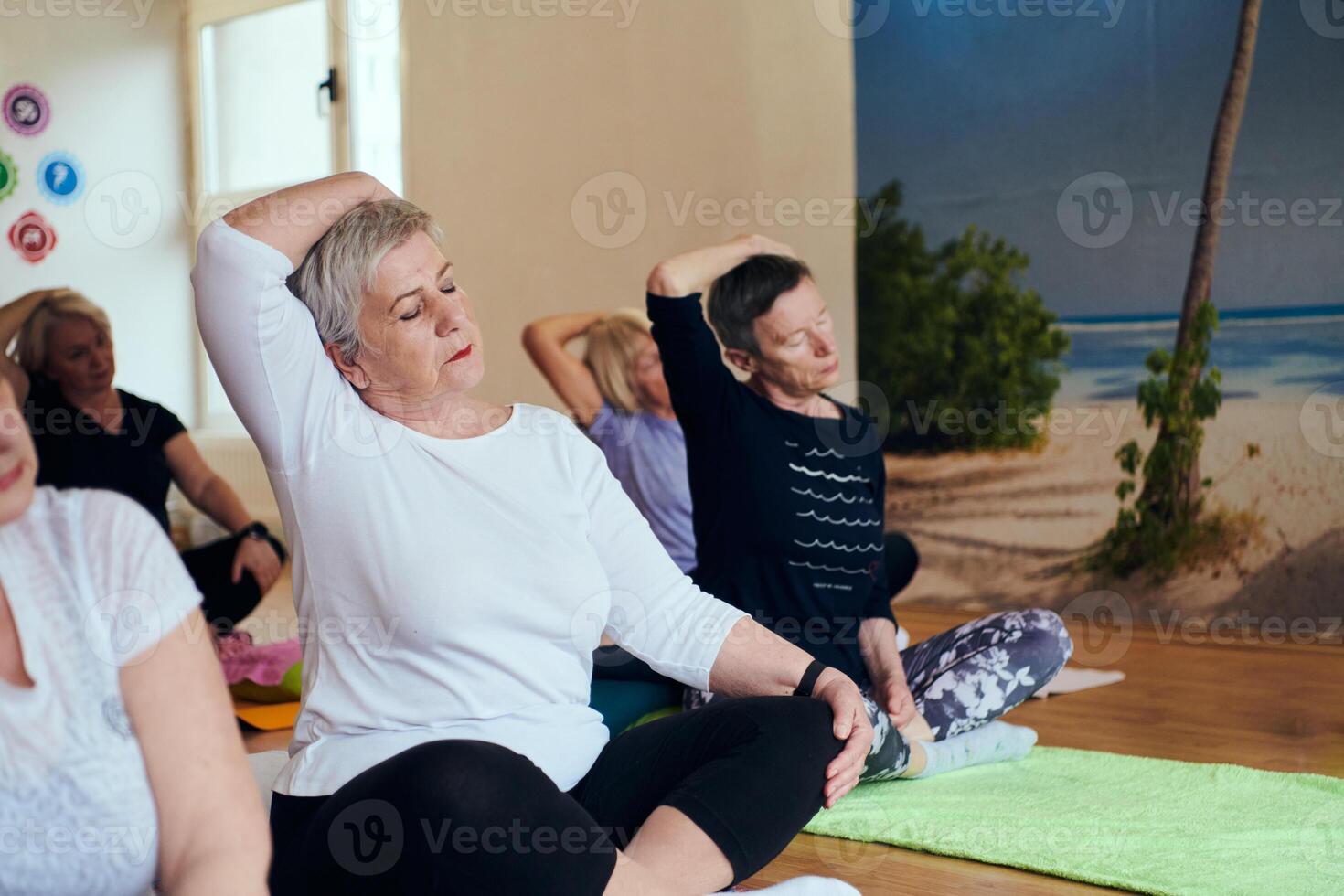 A group of senior women engage in various yoga exercises, including neck, back, and leg stretches, under the guidance of a trainer in a sunlit space, promoting well-being and harmony photo