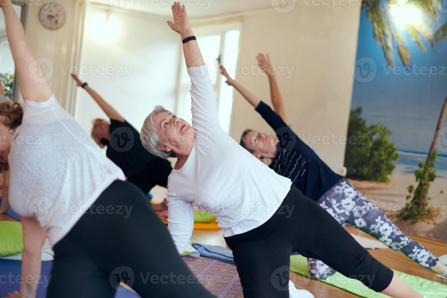 A group of senior women engage in various yoga exercises, including neck, back, and leg stretches, under the guidance of a trainer in a sunlit space, promoting well-being and harmony photo