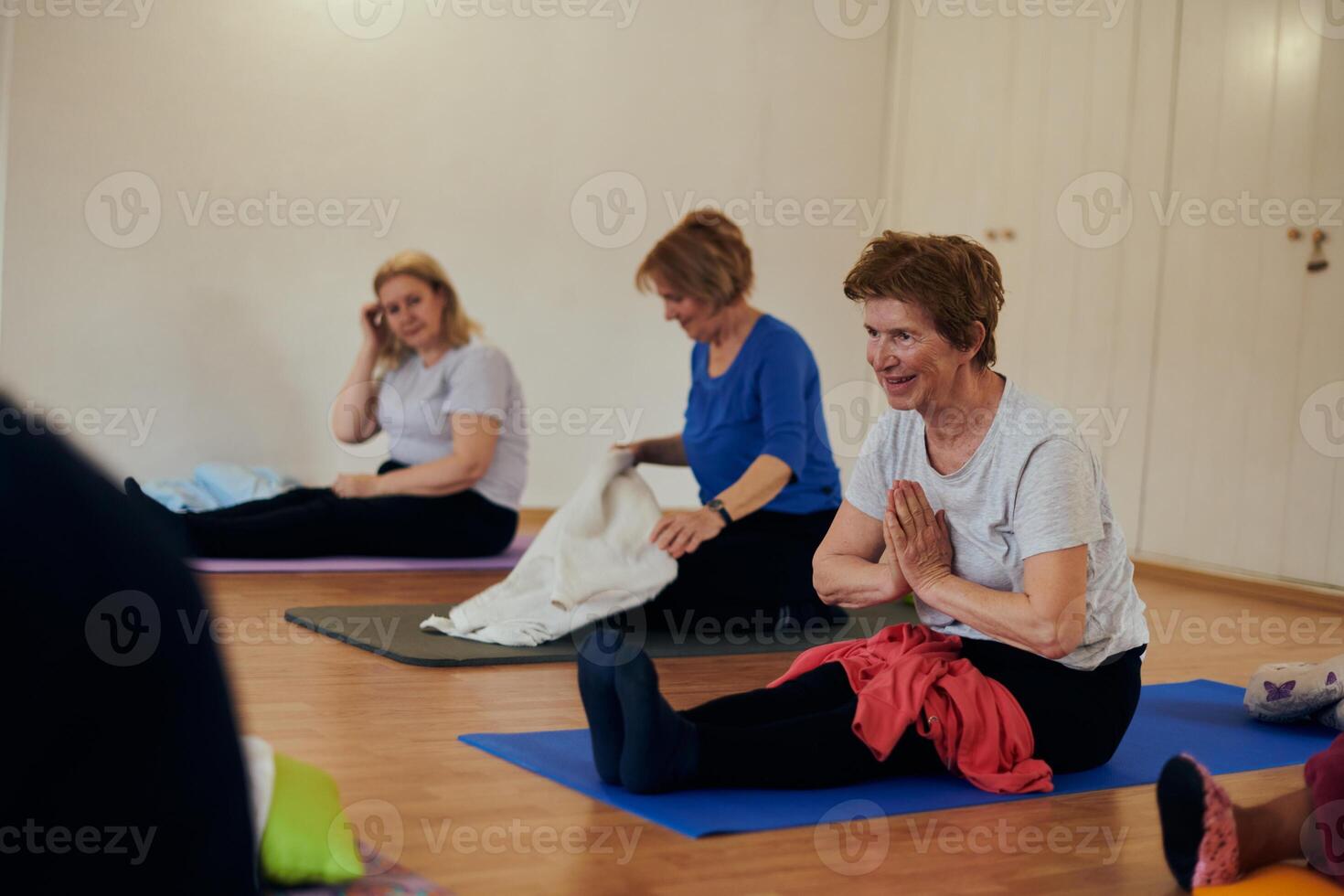 A group of senior women engage in various yoga exercises, including neck, back, and leg stretches, under the guidance of a trainer in a sunlit space, promoting well-being and harmony photo