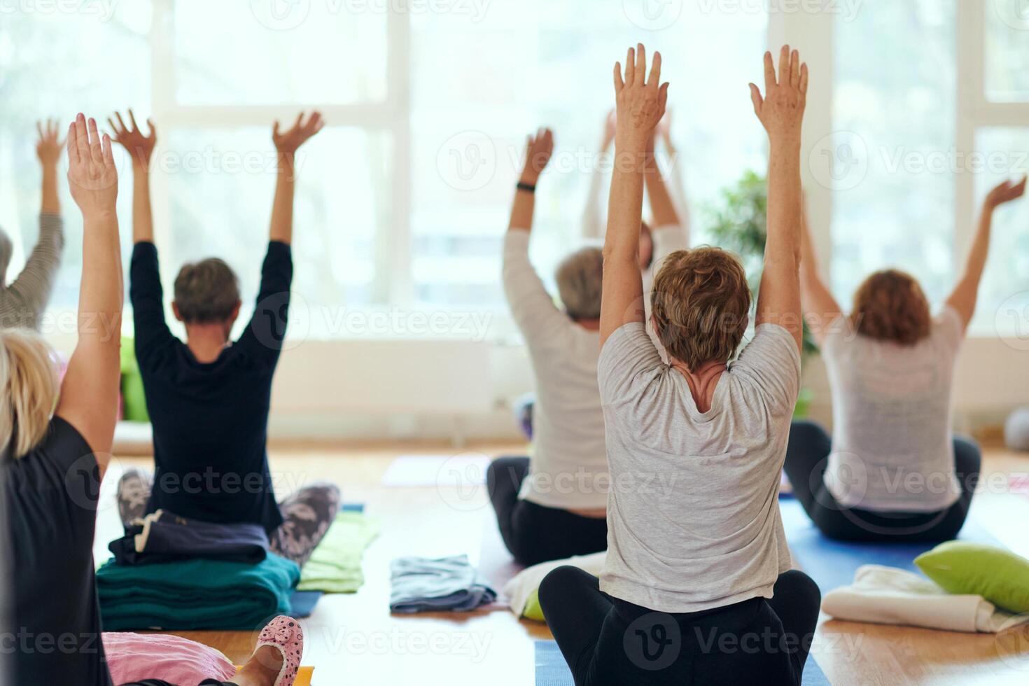 A group of senior women engage in various yoga exercises, including neck, back, and leg stretches, under the guidance of a trainer in a sunlit space, promoting well-being and harmony photo