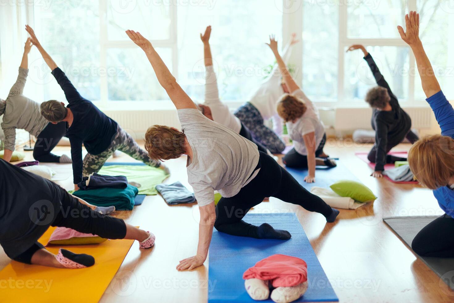 A group of senior women engage in various yoga exercises, including neck, back, and leg stretches, under the guidance of a trainer in a sunlit space, promoting well-being and harmony photo