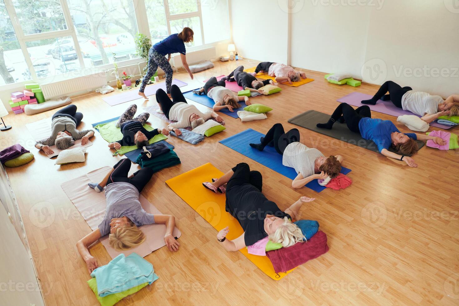 A group of senior women engage in various yoga exercises, including neck, back, and leg stretches, under the guidance of a trainer in a sunlit space, promoting well-being and harmony photo