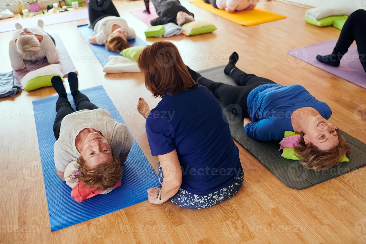 A group of senior women engage in various yoga exercises, including neck, back, and leg stretches, under the guidance of a trainer in a sunlit space, promoting well-being and harmony photo
