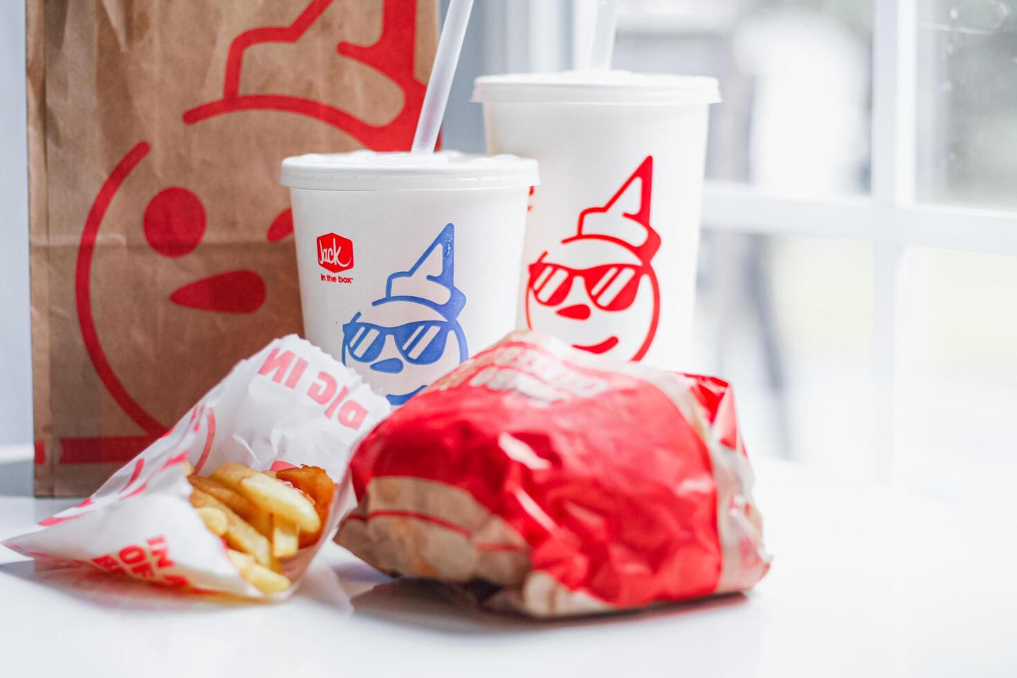 WASHINGTON, USA - August 08, 2024 - A classic fast food spread from Jack in the Box two cups of soda, a bag of fries and a wrapped burger. The cups feature the chain's iconic mascot with sunglasses. photo