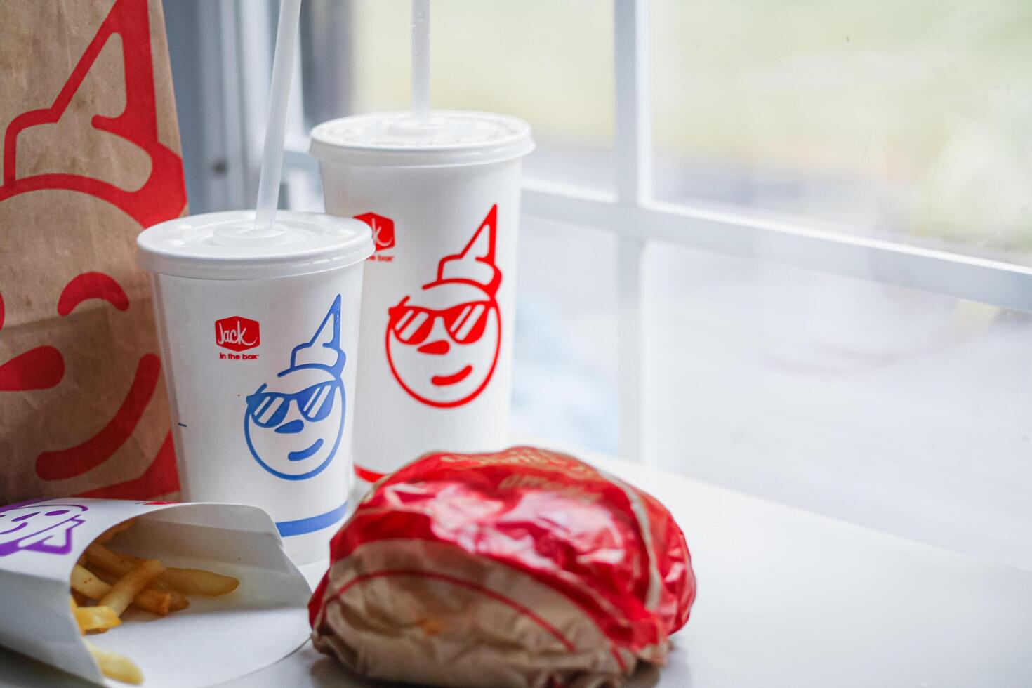 WASHINGTON, USA - August 08, 2024 - A table setting featuring a fast food order from Jack in the Box. The meal includes two white styrofoam cups with red lids and straws, and a wrapped burger. photo