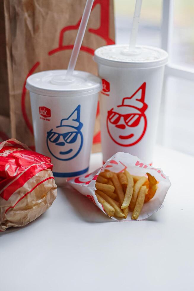 WASHINGTON, USA - August 08, 2024 - A classic fast food spread from Jack in the Box two cups of soda, a bag of fries and a wrapped burger. The cups feature the chain's iconic mascot with sunglasses. photo