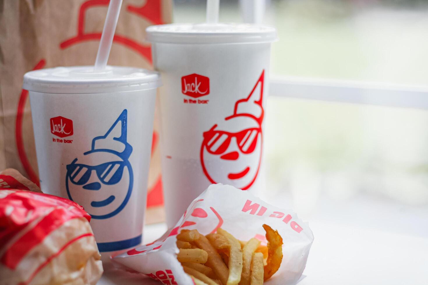 WASHINGTON, USA - August 08, 2024 - A classic fast food spread from Jack in the Box two cups of soda, a bag of fries and a wrapped burger. The cups feature the chain's iconic mascot with sunglasses. photo