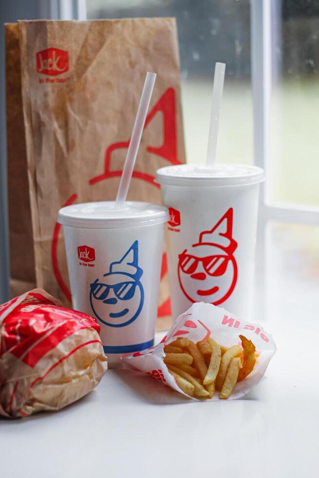 WASHINGTON, USA - August 08, 2024 - A classic fast food spread from Jack in the Box two cups of soda, a bag of fries and a wrapped burger. The cups feature the chain's iconic mascot with sunglasses. photo