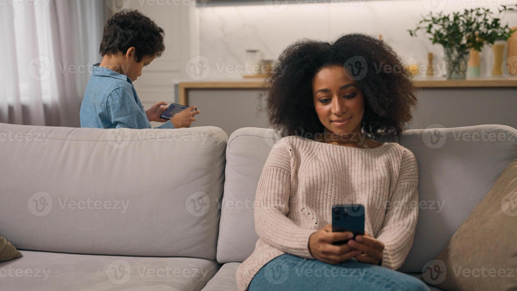 African American family young woman female mother mom chatting online cellphone little child boy kid son playing internet game overuse problem ignoring each other indoors home. Mobile phone addiction photo