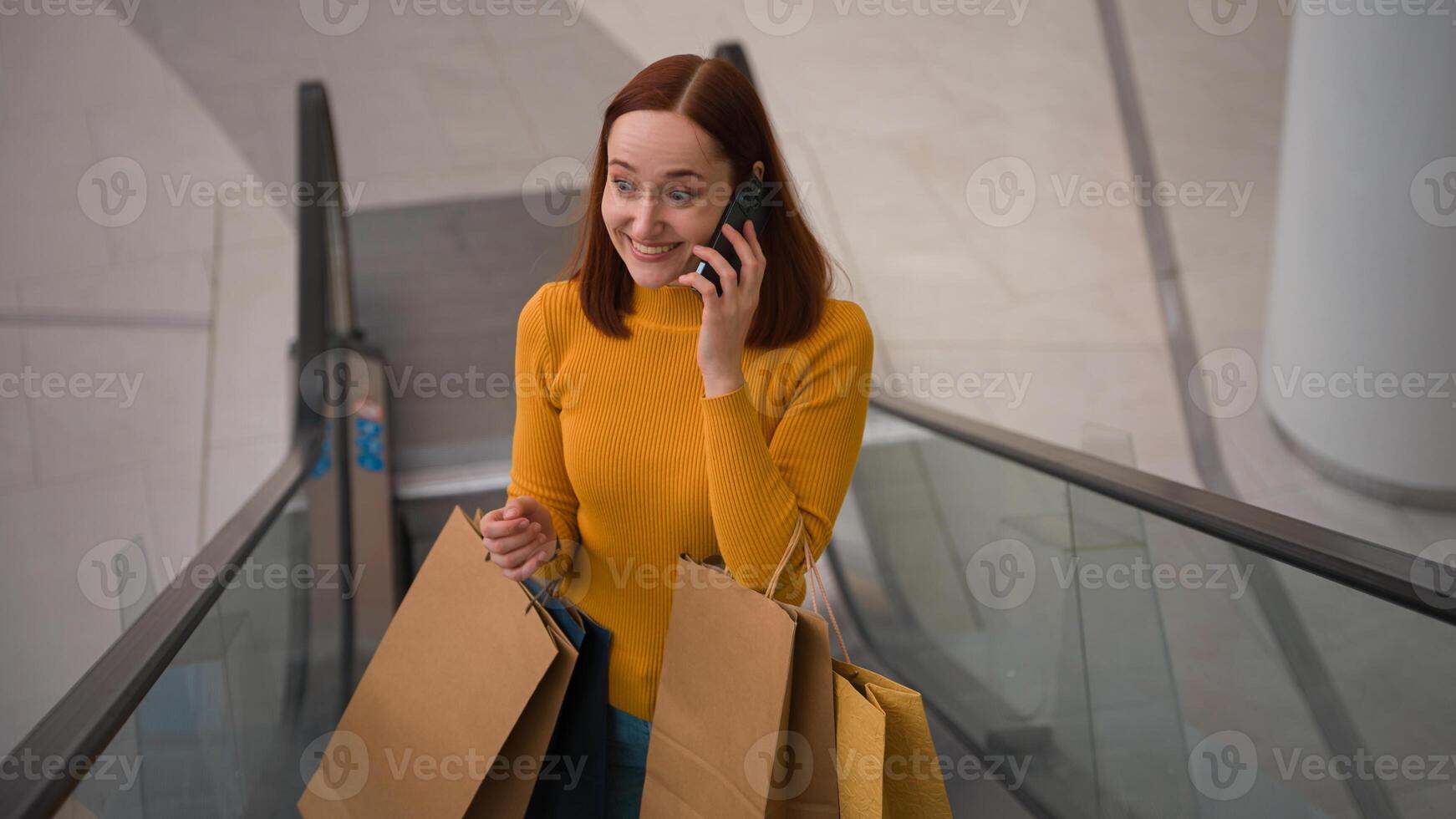 Happy amazed Caucasian girl woman female shopper client customer riding escalator hold paper shop bags purchases talking mobile cell phone call discuss shopping sales discount Black Friday sale indoor photo