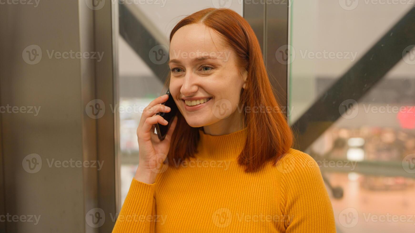 Portrait smiling successful Caucasian red haired girl woman client businesswoman female talk answer speaking mobile cell phone device call moving down indoors glass modern elevator shopping mall store photo