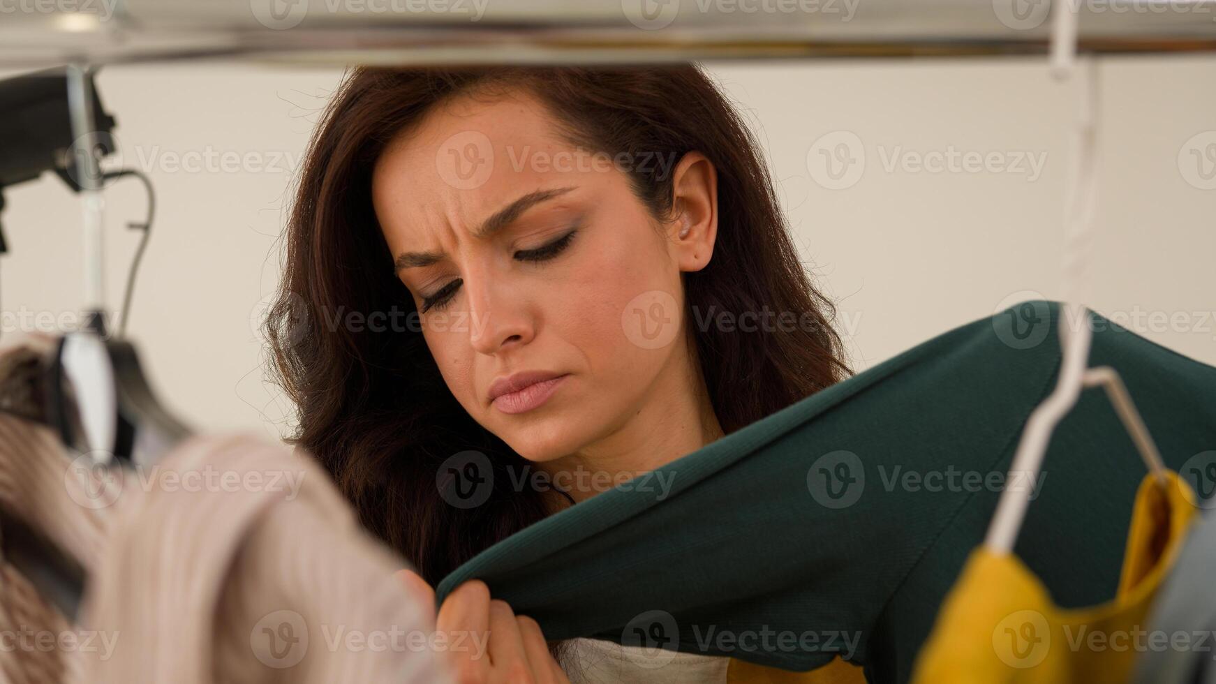 Dissatisfied irritated Caucasian woman vlogger model influencer frown looking choosing touch clothes hangers. Popular female blogger girl picking out shirt for filming advertisement in studio photo