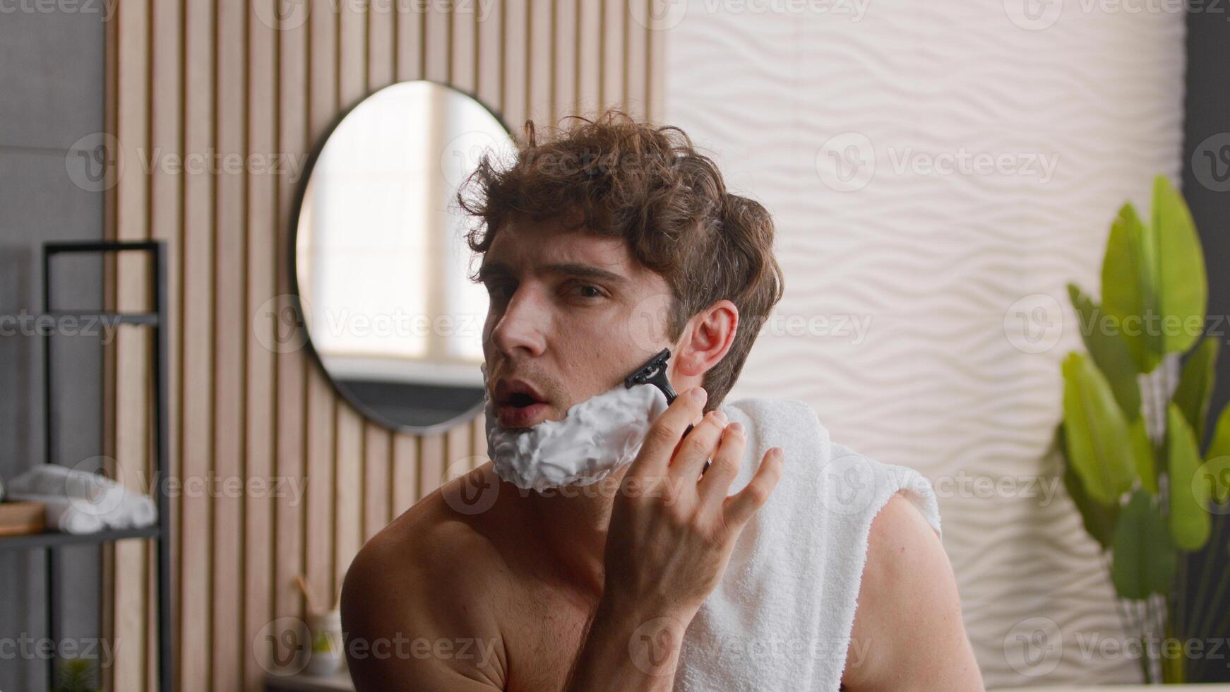 Close up Caucasian bearded male funny man guy looking at camera beauty routine morning hygiene procedure bathroom apply smear face white foam facial shaving gel cosmetic prepare for beard shave bath photo