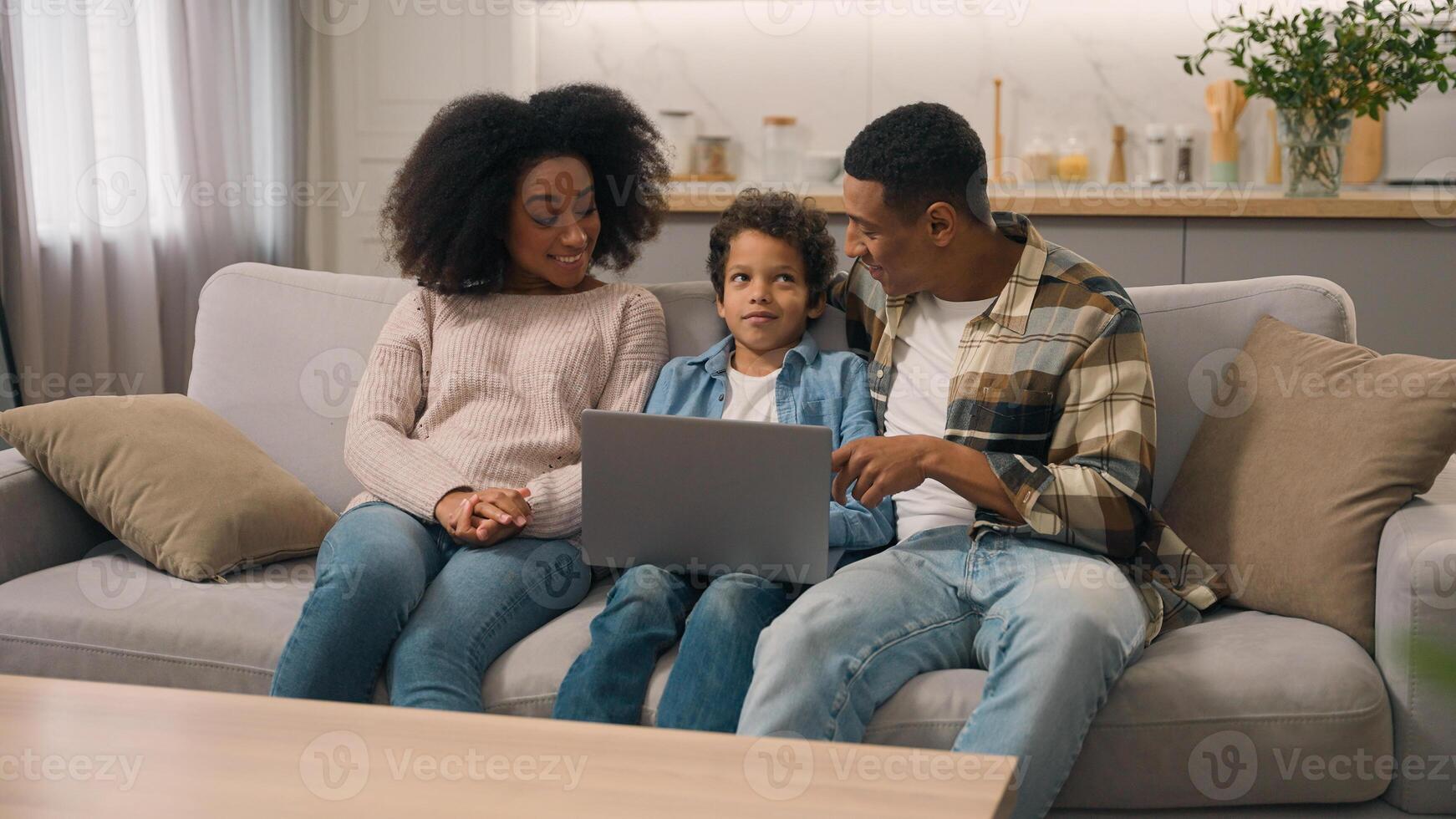 Happy married woman female mom male dad man African American family couple talking explain notebook pc software use internet app little boy child son kid living room couch. Parental control technology photo