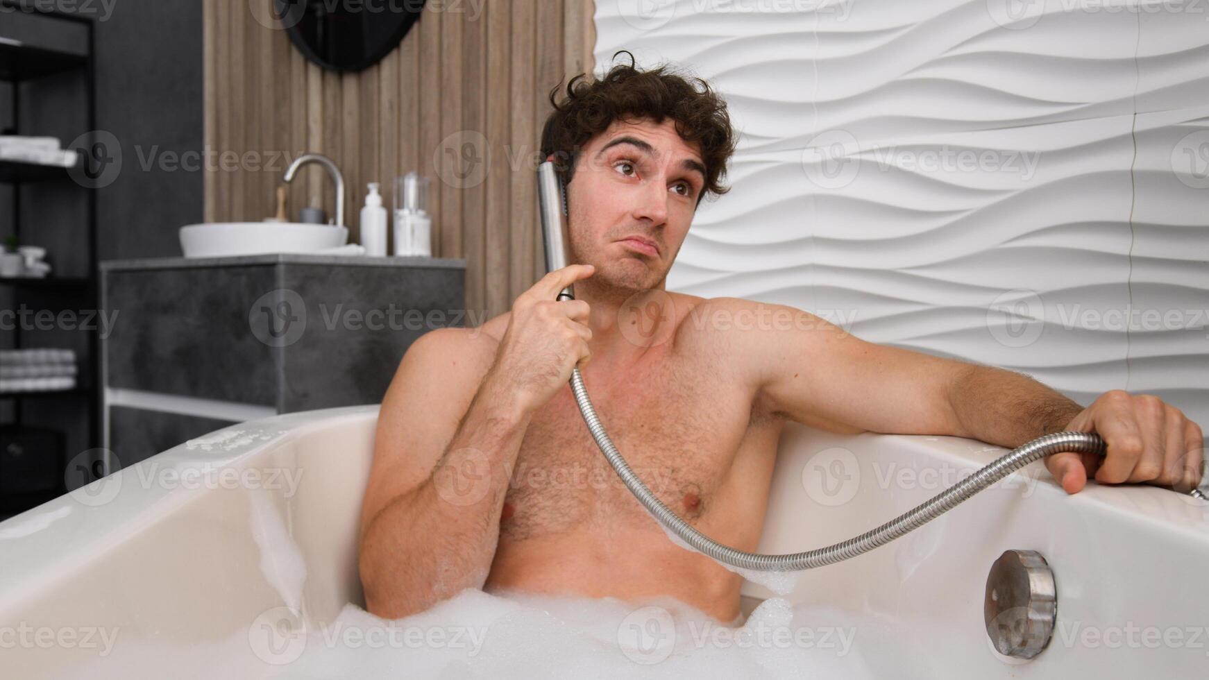 Caucasian man male guy relaxing washing in bath with soapy foam in bathtub speak talking in faucet like telephone mobile phone pretend talk speaking smartphone call silly funny humorous in bathroom photo