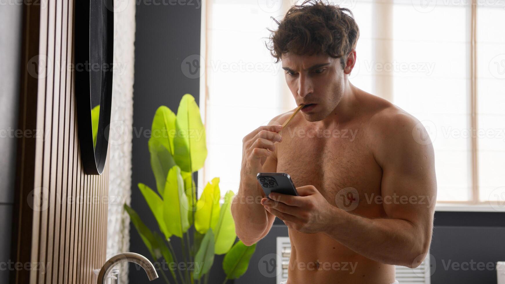 Young Caucasian man brushing teeth in bathroom morning hygiene procedure shocked guy looking in smartphone read bad news failure lost problem oral mouth daily procedure tooth care mobile phone upset photo