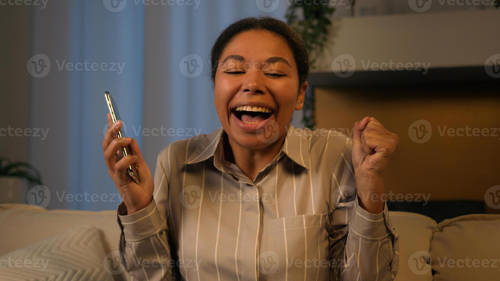 Excited African American woman win online prize in mobile app happy ethnic businesswoman adult female winner shout scream celebrate victory phone lottery money achievement funny dance at evening home photo