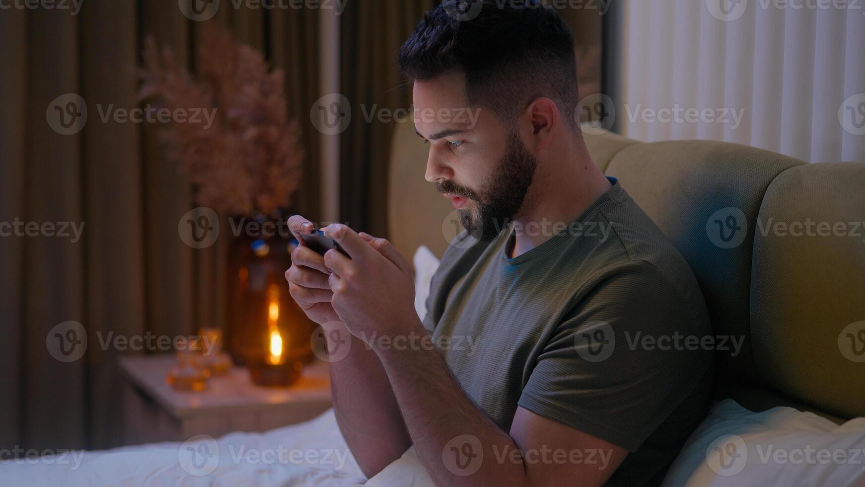 Caucasian man using mobile phone smartphone telephone gadget online virtual game playing glued stuck addiction male guy evening digital device cyber tapping scrolling connected electronic bedroom bed photo