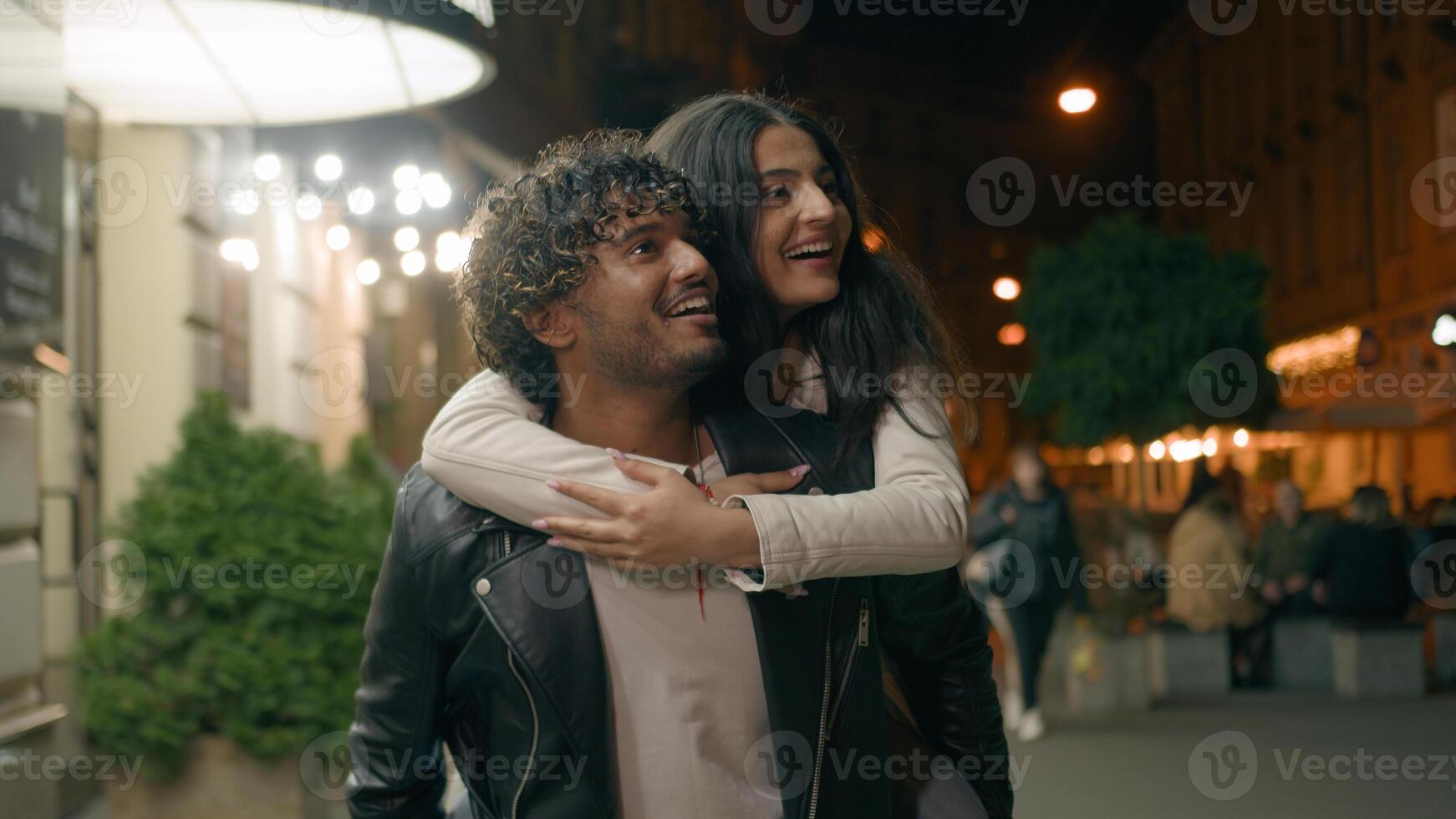 Indian Arabian couple love guy holding girl on back piggyback woman man boyfriend girlfriend walking night evening city looking at beautiful urban architecture pointing excited tourist travel dating photo