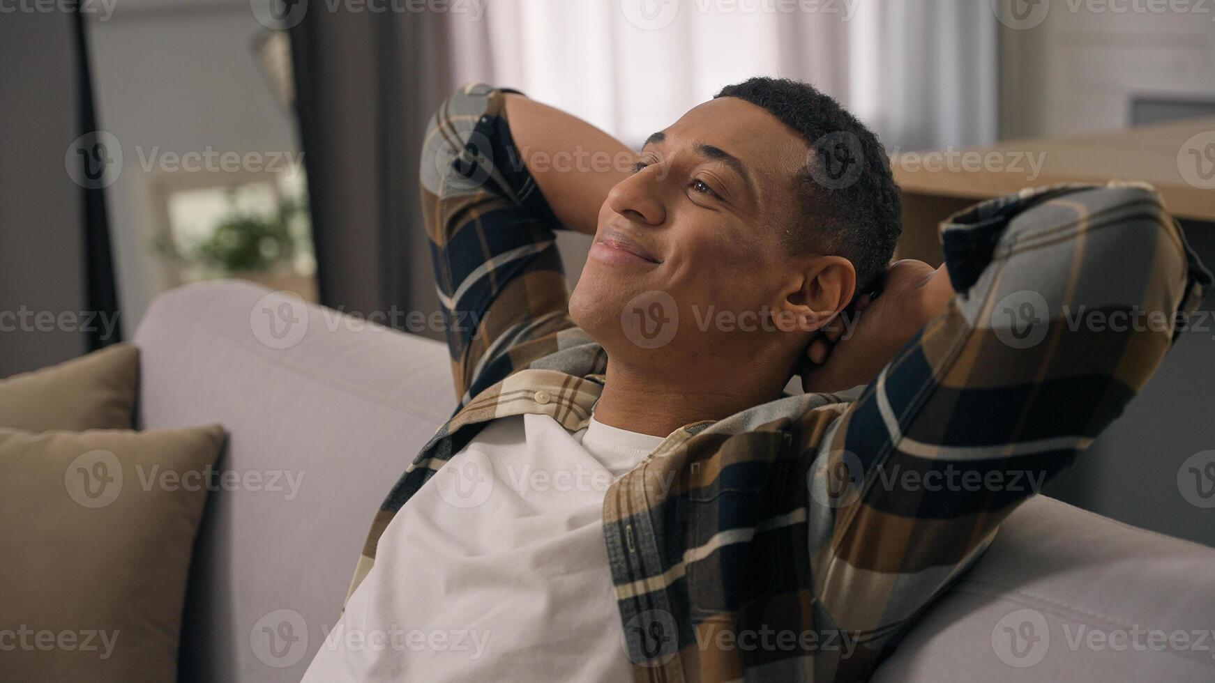 Smiling happy inspired dreaming African American man biracial guy ethnic male relax at home sofa comfortable couch leaning back hands behind head rest apartment weekend resting dream nap closed eyes photo