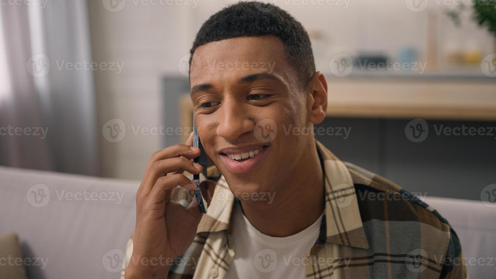 Smiling African American ethnic man carefree guy smile male talking call smartphone talk speaking mobile phone speak communicate laughing conversation gadget at home couch sofa chatting glad happy photo