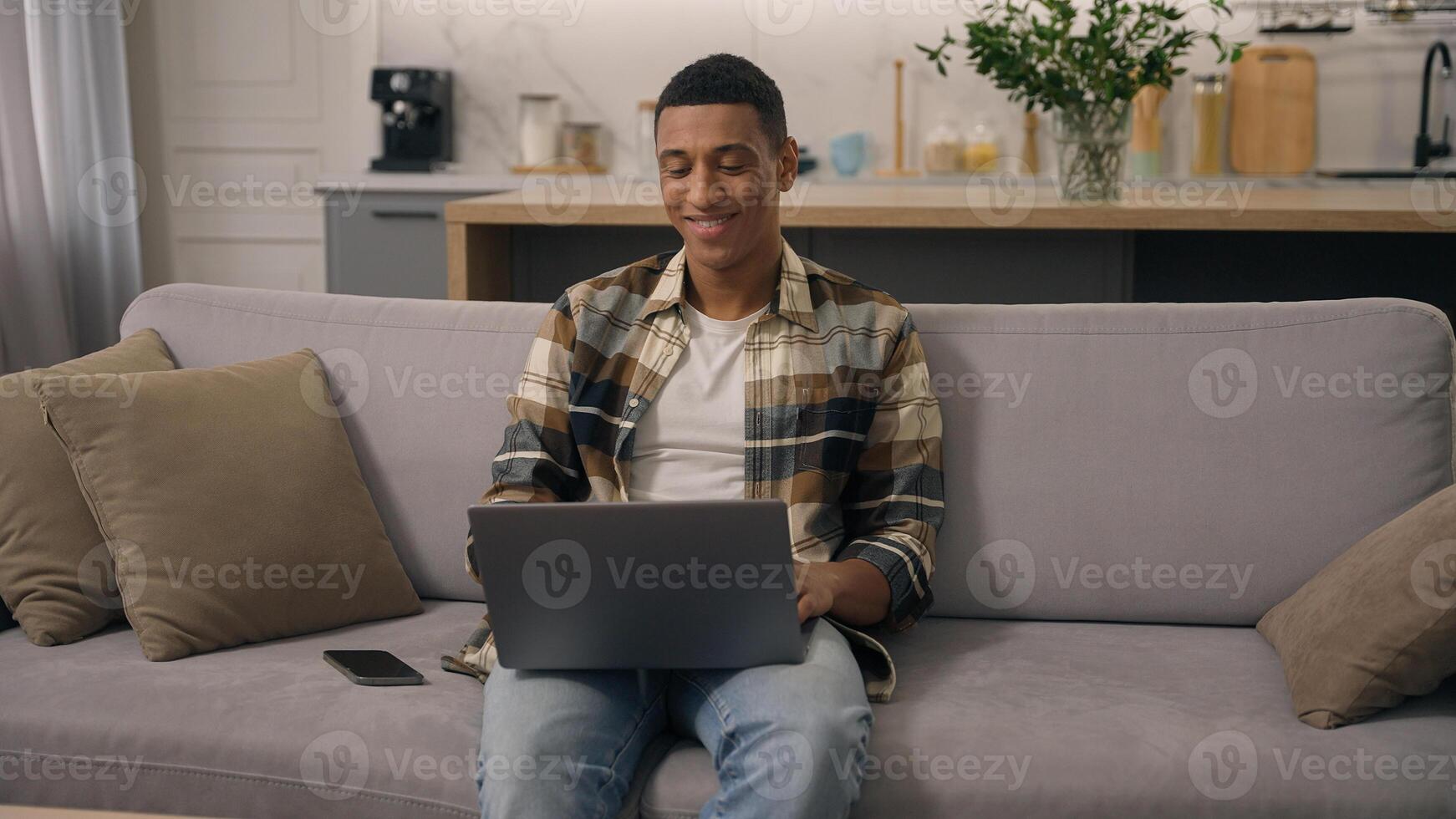 Happy smiling African American ethnic man working at home distant online work studying at sofa guy laptop using computer at couch male using pc playing internet game use app shopping chatting photo