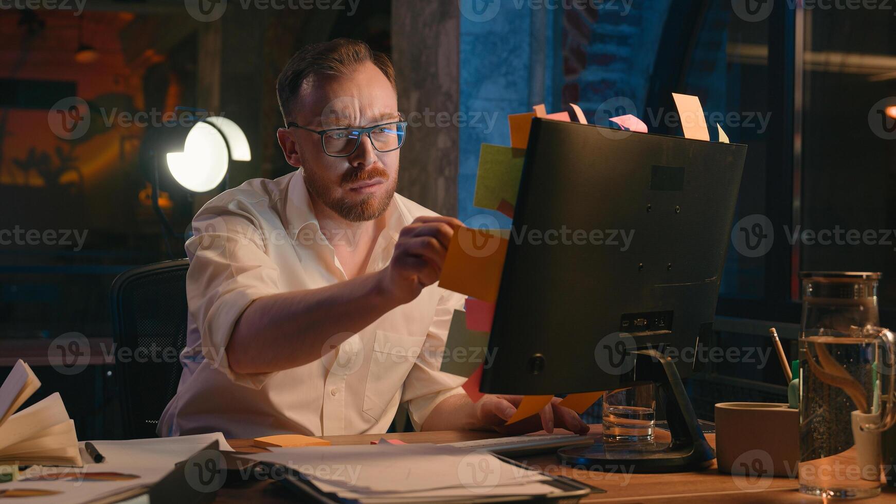 Caucasian man business guy businessman male in dark night evening office work reading check information on stickers throw away dissatisfied read sticky paper notes on computer monitor screen deadline photo