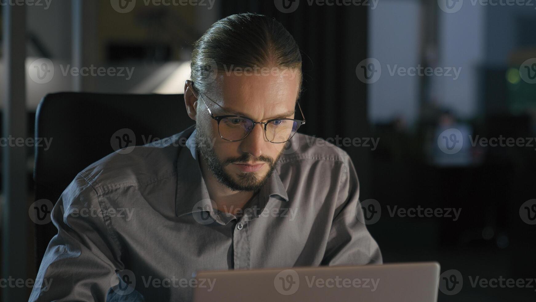 Overtime working businessman manager man at night evening dark office work with laptop smiling toothy happy business man worker leader employer with computer job at table workplace looking at camera photo