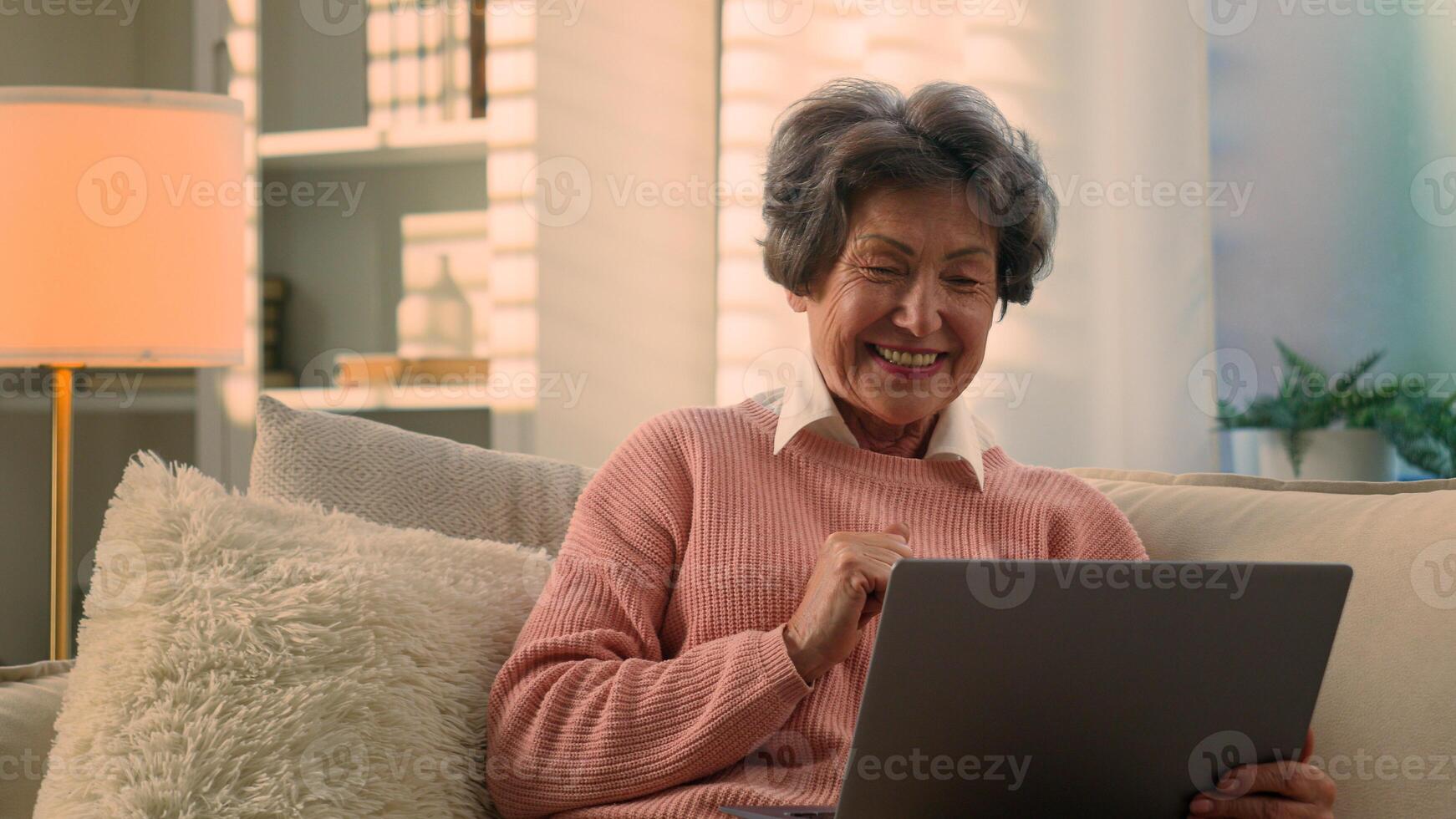 Carefree joyful smiling retired Caucasian happy cheerful old woman senior lady mature retirement elderly female smile talking pleasant conversation online chat call with laptop at home couch photo