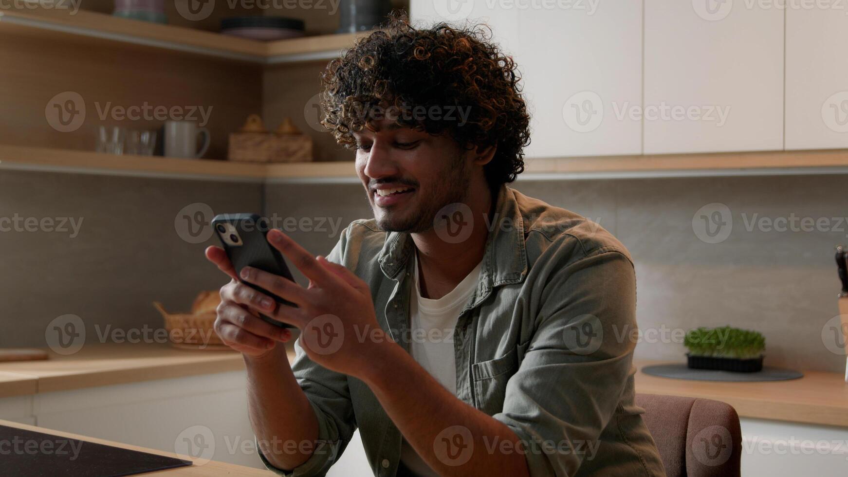 Satisfied Indian male win with phone winning financial prize reward goal achievement with mobile app betting service online victory. Arabian happy man celebrating smartphone success at home kitchen photo
