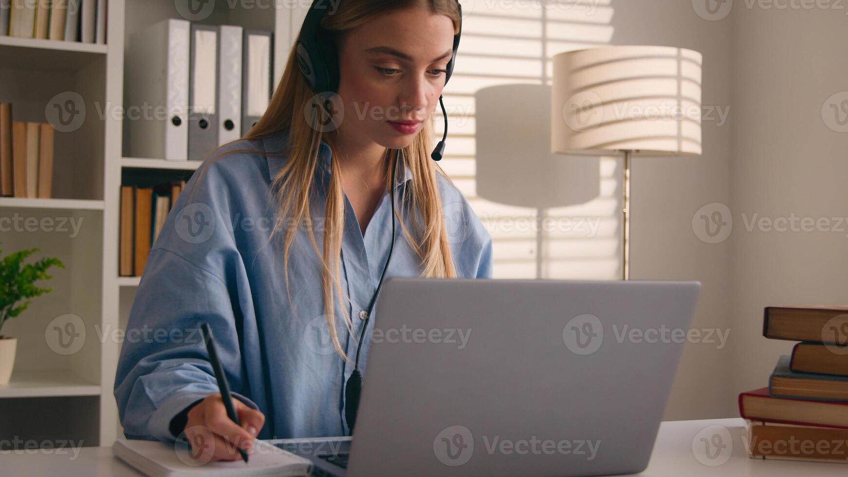 Caucasian woman in headset conference calling on laptop online teacher studying e-learning from home library with books writing notes write homework student girl learning computer distance education photo