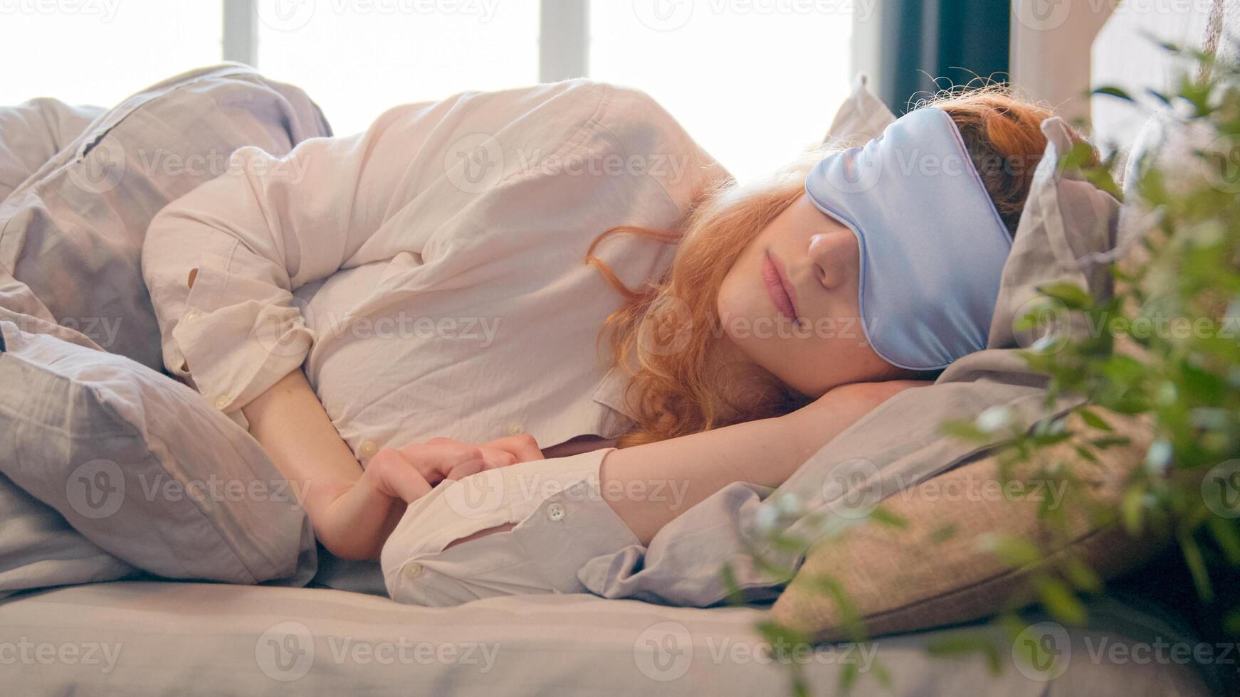 Lazy Caucasian girl with blindfold in bed in morning woman sleeping napping during daytime sleepy female waking up lying in comfortable bed peeks out from under sleep mask opens eye fall asleep again photo