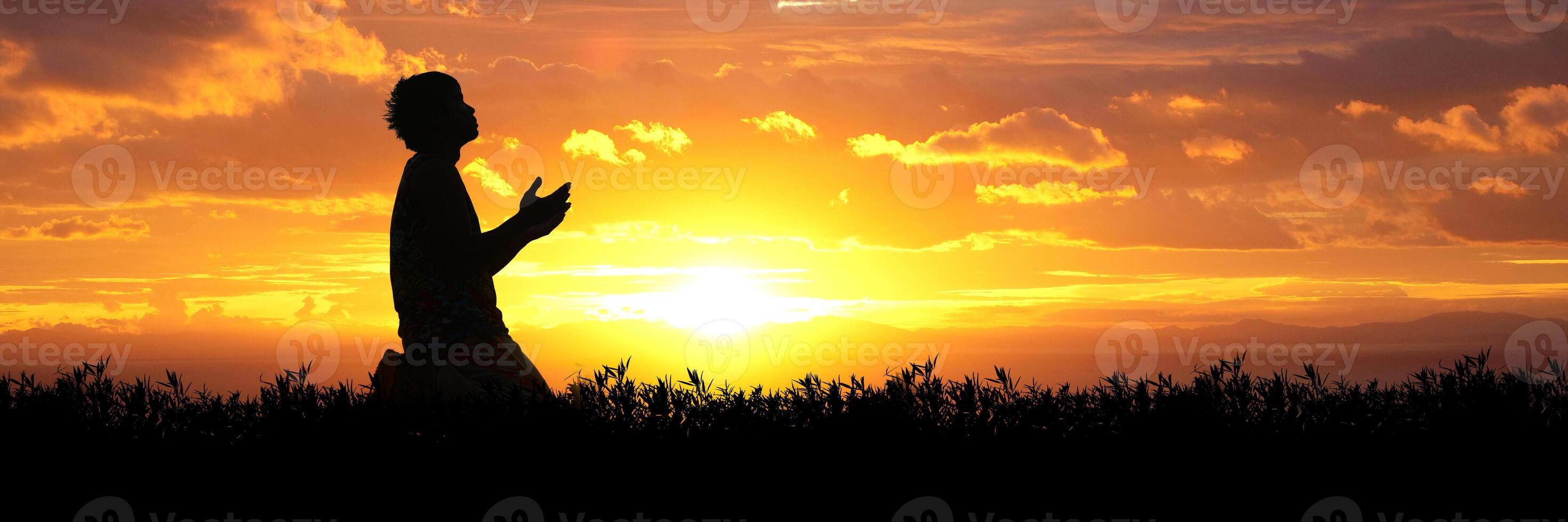 Silhouette of a Person Praying at Sunset with a Vibrant Orange and Yellow Sky in the Background photo