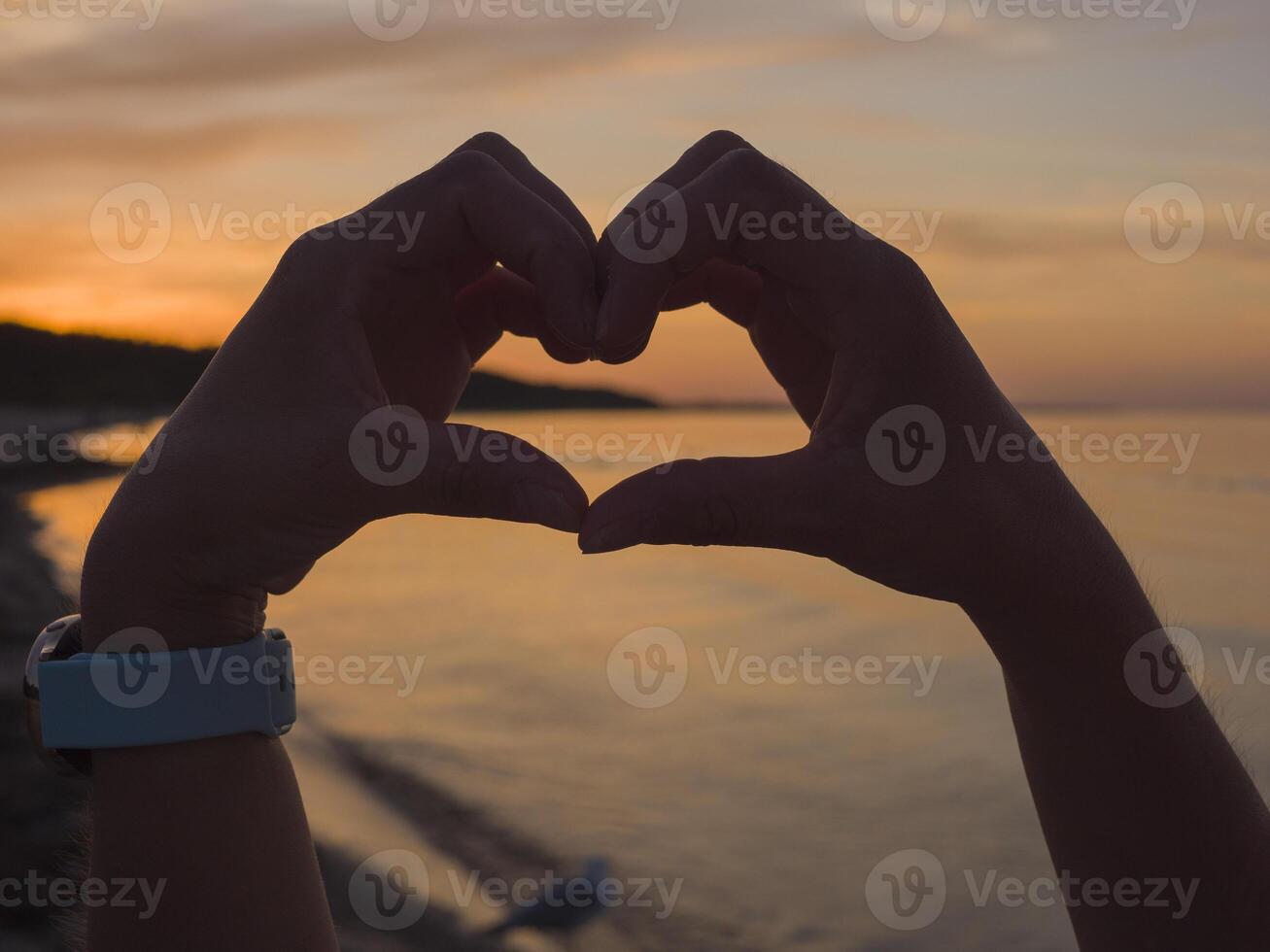 Hands form a heart-shaped silhouette against a sunset, where the suns glow softly illuminates the heart-shaped outline. This heart-shaped gesture resonates with warmth and love as day turns to night. photo