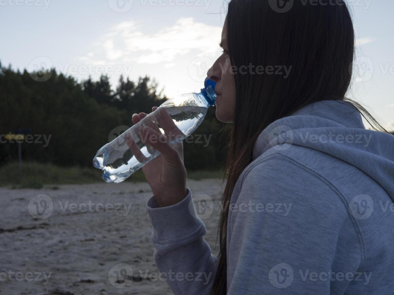 A young brunette woman sits by the sea, drinking mineral water from a plastic bottle. As she relaxes on the shore, the cool taste of mineral water provides a refreshing escape from the world. photo