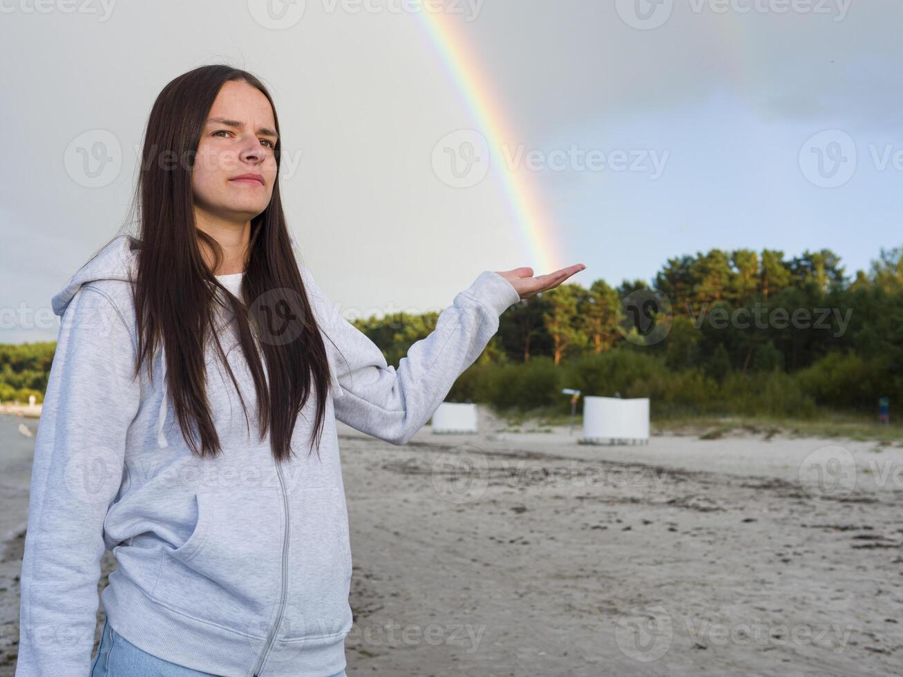 On the beach, a Woman Young Brunette reaches out, catching a Rainbow in her hand. The delicate hues of the Rainbow dance against backdrop of waves, as she gazes at Rainbow's shimmering light. photo