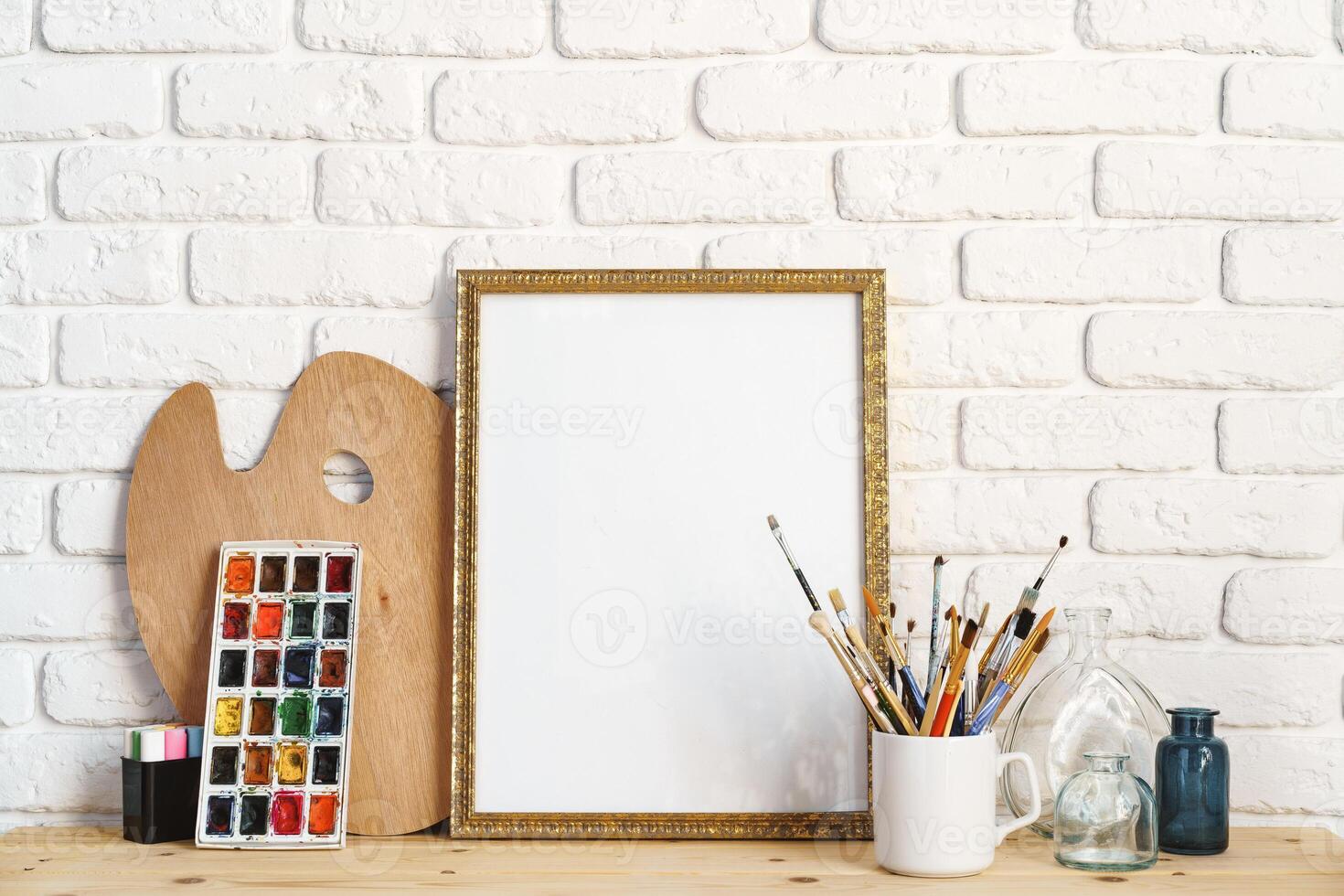 Photo frame as easel with artist's tools on wooden table against white brick wall