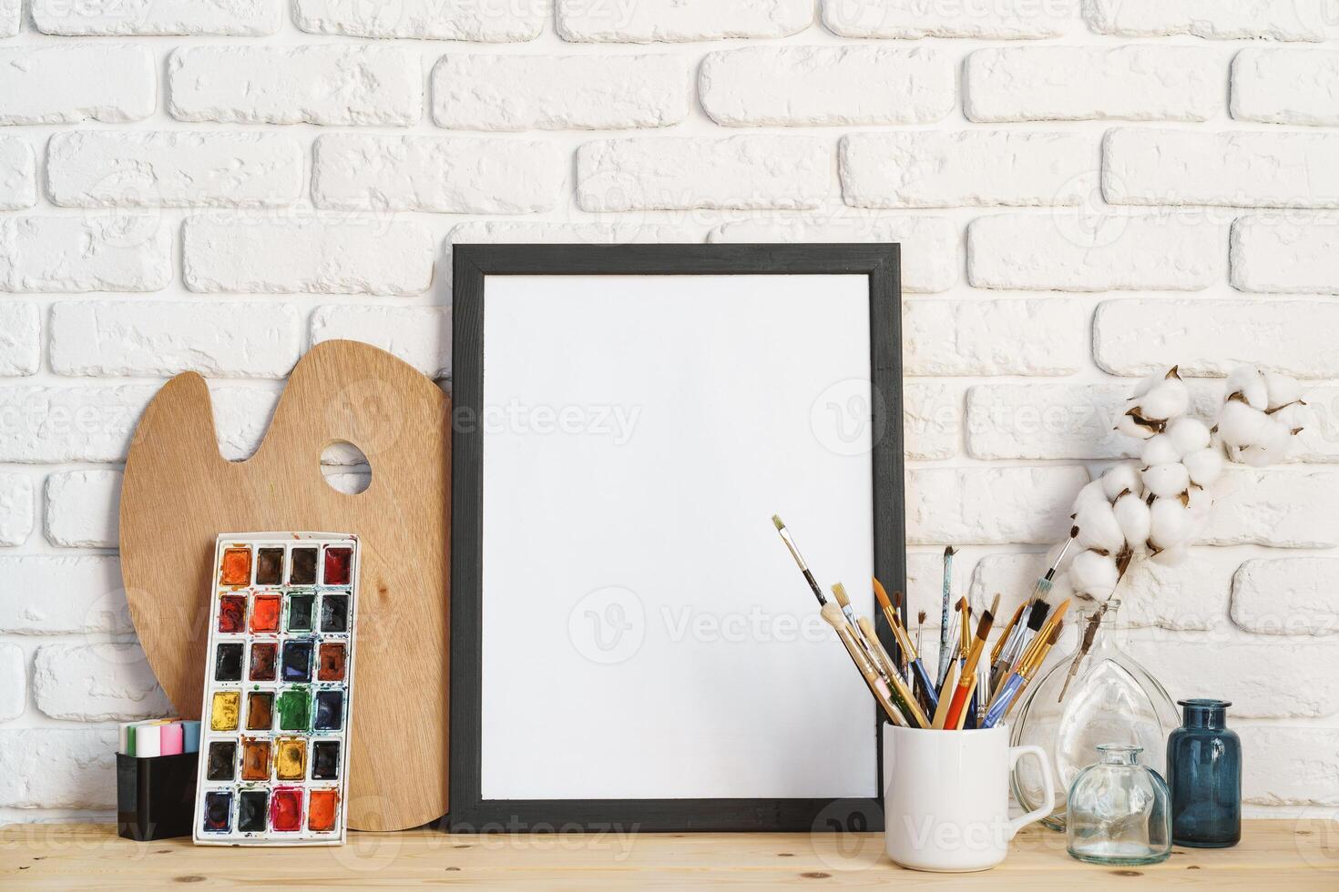 Photo frame as easel with artist's tools on wooden table against white brick wall