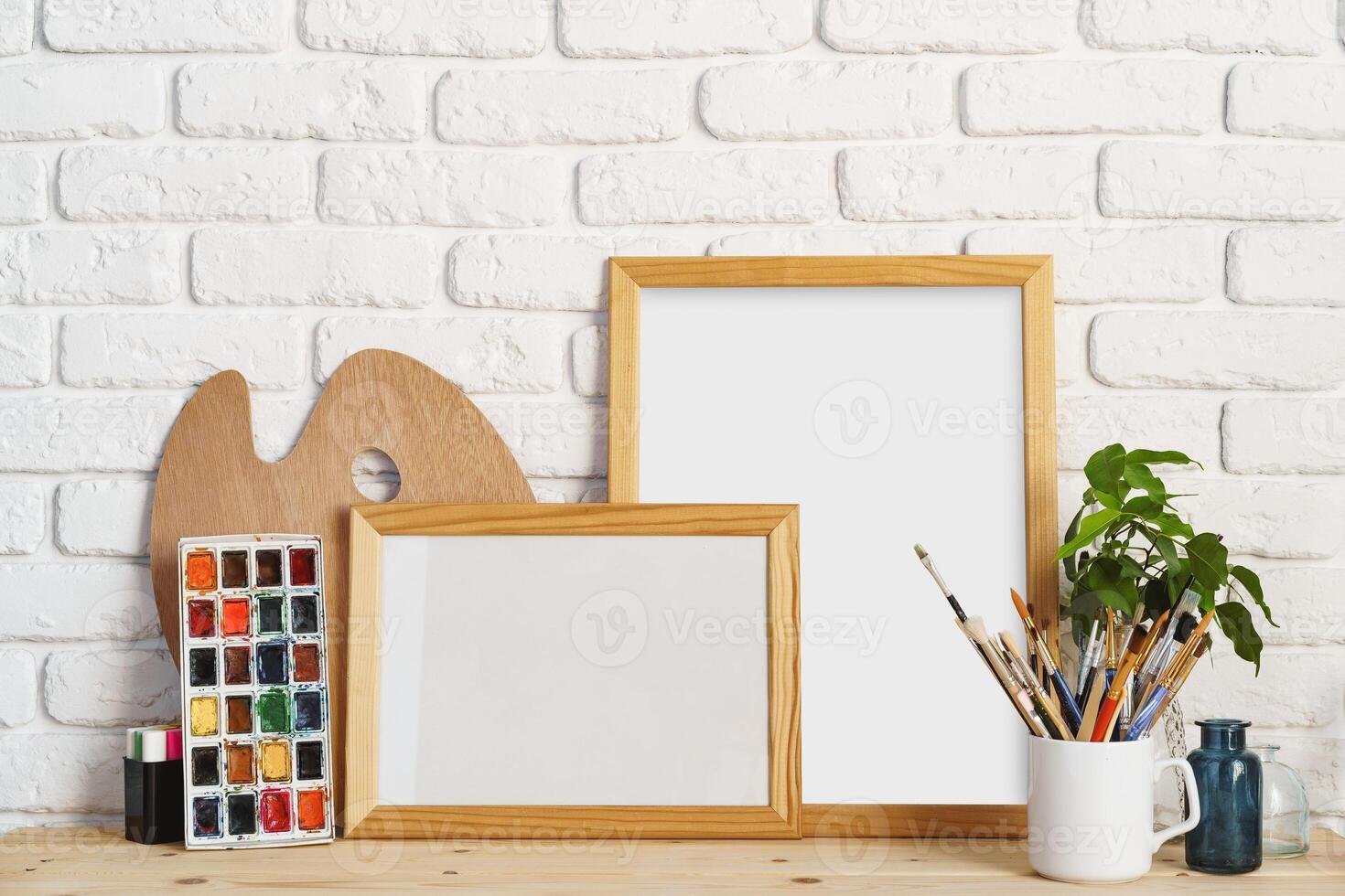 Photo frame as easel with artist's tools on wooden table against white brick wall